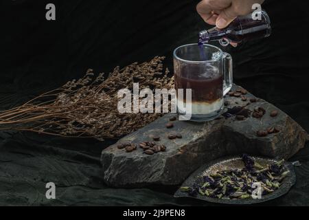 Gießen Sie Butterfly-Erbsensaft (Anchan) aus einer kleinen Glasflasche in das Glas Espresso-Kaffee und frische Milch am Boden mit gerösteten Kaffeebohnen Stockfoto