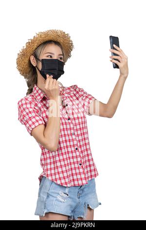 Isoliertes Halbbild einer Touristenin mit rotem kariertem Hemd, kurzen Jeans und Strohhut, die Selfie macht, während die Hand ein Mini-Herzzeichen zeigt. Stockfoto