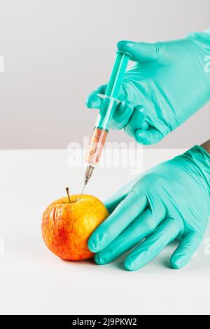 Eine Hand in einem medizinischen Handschuh führt eine Spritze in den Apfel ein. Schädliche Lebensmittelzusatzstoffe. GVO-Konzept Stockfoto