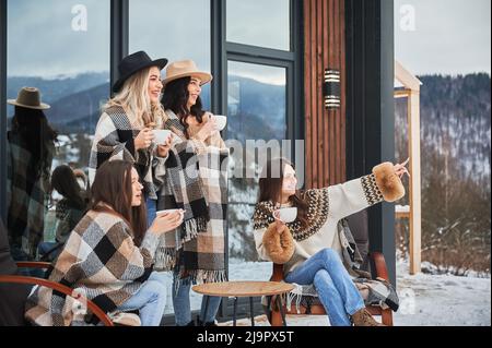 Junge Frauen genießen Winterwochenenden auf der Terrasse des modernen Scheunenhauses. Vier Mädchen, die Spaß haben, auf Stühlen sitzen und heißen Tee trinken. Stockfoto