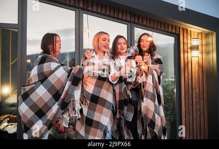 Junge Frauen genießen die Winterwochenenden auf der Terrasse des modernen Barnhouse. Vier Mädchen in Plaids trinken Wein und feiern mit Wunderkerzen am Abend. Stockfoto