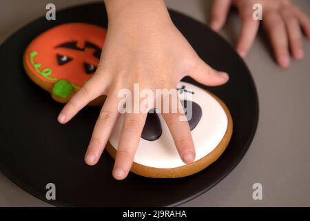 Die Hand des Mädchens greift nach hausgemachten Halloween-Keksen mit bemalten Gesichtern Stockfoto