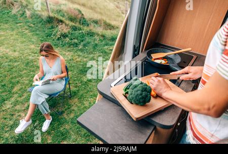 Mann kocht Gemüse in einem Wohnmobil, während Frau draußen liest Stockfoto