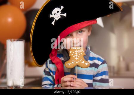 Ein als Pirat gekleideter Junge isst Lebkuchen als Halloween-Skelett, trinkt Milch Stockfoto