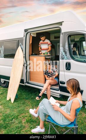 Freunde, die in einem Wohnmobil unterwegs sind. Mann kocht, Frau liest und Frau schaut mobil Stockfoto