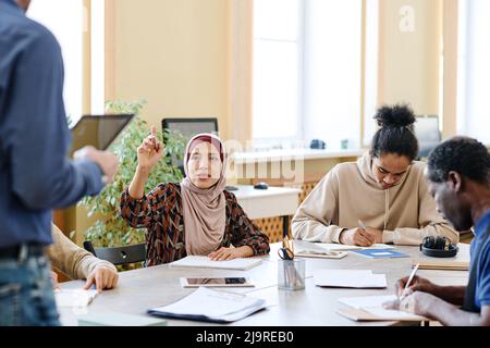 Muslimische Frau, die Sprachunterricht für Immigranten nimmt und die Hand hebt, um dem Lehrer Fragen über die Aufgabe zu stellen Stockfoto