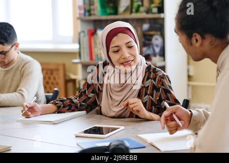 Muslimische Frau mit Hijab, die am Tisch in der Bibliothek sitzt und während des Unterrichts für Einwanderer in das Notizbuch ihres Klassenkameraden schaut Stockfoto