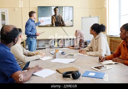 Eine Gruppe von multiethnischen Einwanderern, die während des Englischuntergangs am Tisch sitzen und sich ein Bildungsvideo ansehen Stockfoto