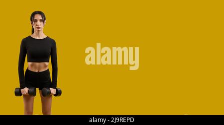Vorderansicht der Brünette Mädchen trainieren, trainieren, Training mit Hanteln. Sportliche Frauen stehen, betrachten die Kamera und tragen Sportkleidung. Isoliert auf gelbem Studiohintergrund. Stockfoto