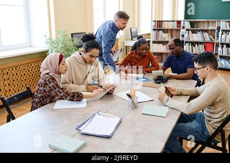 Reifer kaukasischer Englischlehrer hilft eingewanderten Schülern dabei, während des Unterrichts in der Bibliothek Aufgaben zu erledigen Stockfoto