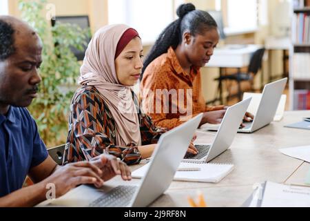 Schwarze und nahöstliche Einwanderer, die an einem Englischkurs teilnehmen, sitzen am Tisch und schreiben etwas auf Laptops Stockfoto