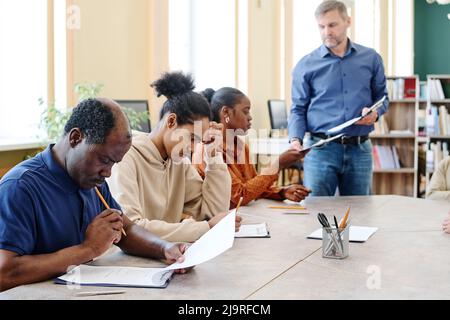 Gruppe moderner schwarzer Einwanderer, die an Kursen am Tisch sitzen und den englischen Grammatiktest machen Stockfoto