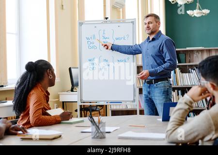 Reifer kaukasischer Mann, der in der Schule für Migranten arbeitet und die Grammatikregeln für englische Fragen erklärt Stockfoto