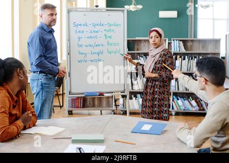 Eine Gruppe von multiethnischen Einwanderern, die in der Schule Englisch lernen, macht Aufgaben mit unregelmäßigen Verben zusammen Stockfoto