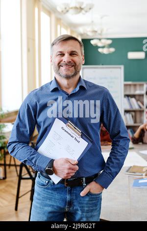 Vertikale mittlere Aufnahme von freudigen reifen kaukasischen Mann Unterricht Englisch Sprache halten Zwischenablage mit Grammatiktests lächeln an der Kamera Stockfoto