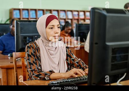 Porträt einer nachdenklichen muslimischen Frau mit Hijab, die in der Universitätsbibliothek sitzt und etwas auf dem Desktop-Computer schreibt Stockfoto