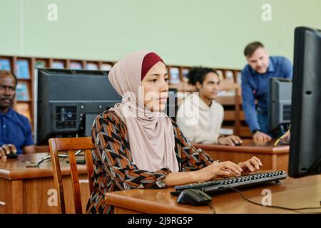 Porträt einer modernen muslimischen Frau mit Hijab, die in der Universitätsbibliothek sitzt und auf einem Desktop-Computer arbeitet Stockfoto