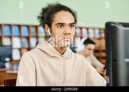 Moderner Typ der Z-Generation, der schnurlose Ohrhörer trägt und am Schreibtisch in der Universitätsbibliothek sitzt und am Desktop-Computer arbeitet und Musik hört Stockfoto