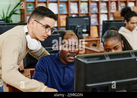 Junger Mann aus dem Nahen Osten und eine schwarze Frau helfen dem reifen Mann, in der Universitätsbibliothek etwas im Internet zu finden Stockfoto