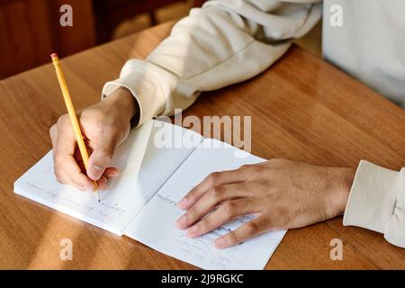 Mittlere Nahaufnahme eines unerkennbaren jungen Mannes, der an der Schule am Schreibtisch sitzt und etwas in einem Notizbuch schreibt Stockfoto