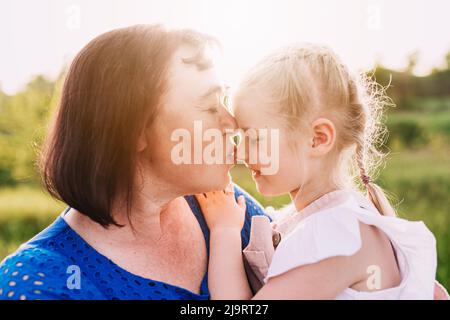 Großmutter hielt Enkelin auf ihren Armen und küsste die Nase. Glückliche Kindheit, Familienliebe Konzept. Unterstützung und Zweisamkeit. Zwei Personen im Freien mit Sonnenlicht Hintergrund. Hochwertige Fotos Stockfoto