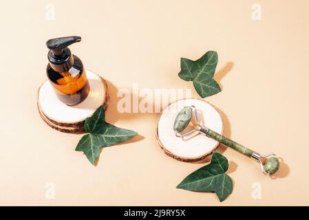 Braune Glasflasche und Jade-Gesichtswalze auf Holzständer mit grünen Efeu-Blättern auf neutralem beigen Hintergrund. Naturkosmetik, Spa und Wellnes Stockfoto