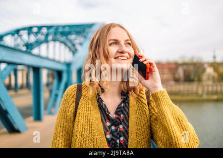 Attraktive junge kaukasische blonde Haare 30s Frau in lässigem Kleid lächelnd, ruft per Smartphone auf der Spring City Street. Stockfoto