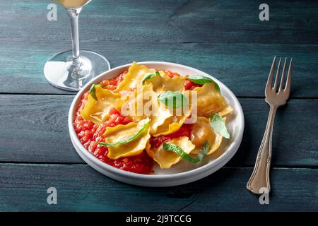Ravioli mit Tomatensauce und frischen Basilikumblättern auf einem Teller, italienisches Abendessen, auf einem dunklen Holztisch mit einem Glas Wein Stockfoto