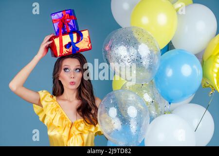 Glücklich überrascht und erstaunt Frau hält eine Geschenkbox auf blauem Hintergrund und Helium gelb, blau und weiß Ballons.Boxen von Geschenken in den Händen eines Stockfoto