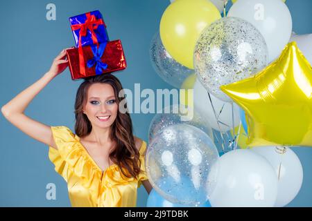 Glücklich lächelnde Frau hält eine Geschenkbox auf blauem Hintergrund und Heliumgelb, blau und weiß Ballons.Boxen von Geschenken in den Händen eines überraschten Mädchen in Stockfoto