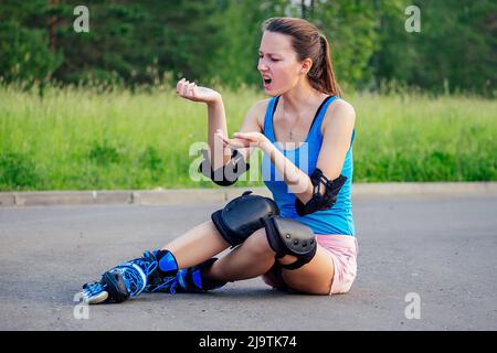 Attraktive junge athletische schlanke Brünette Frau in rosa Shorts und blauem Oberteil mit Schutz Ellenbogenschützern und Kniepolstern auf Rollschuhe sitzen auf dem Stockfoto