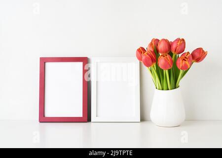 Zwei Bilderrahmen-Mockups und rote Tulpen in einer weißen Vase auf einem weißen Tisch an der Wand. Innenraum, Platz für Text kopieren Stockfoto