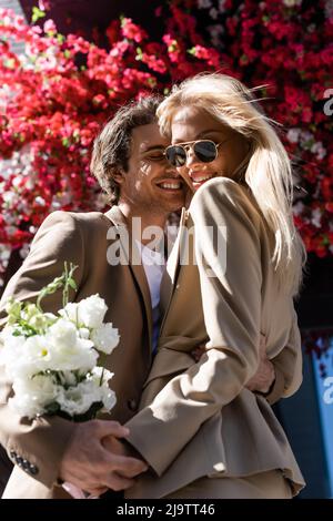 Erfreut Frau in Sonnenbrille hält Strauß in der Nähe glücklichen Mann umarmt sie im Freien Stockfoto