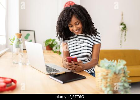 Junge hispanische amerikanerin, die zu Hause ein Mobiltelefon benutzt, während sie am Laptop arbeitet Stockfoto