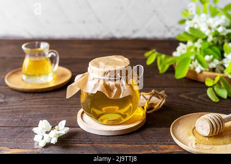 Akazienhonig im Glasgefäß mit Blumen auf Holzgrund zur Seite Stockfoto