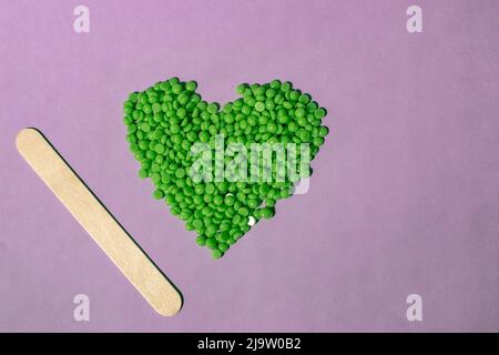 Schöne grüne Wachs-Granulat mit Aloe-Extrakt zur Enthaarung werden auf einem hellvioletten Hintergrund gegossen, in Form eines Herzens. Holzspatel. Epilat Stockfoto