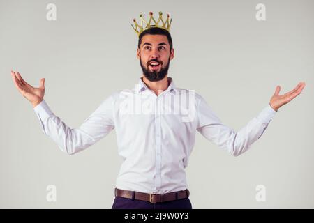 Porträt eines lächelnden bärtigen Geschäftsmanns mit einer goldenen Krone auf dem Kopf auf weißem Hintergrund, aufgenommen im Studio Stockfoto