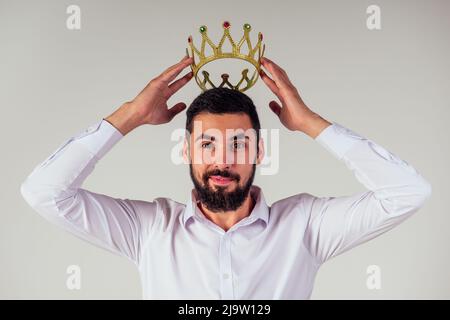Porträt eines lächelnden bärtigen Geschäftsmanns mit einer goldenen Krone auf dem Kopf auf weißem Hintergrund, aufgenommen im Studio Stockfoto