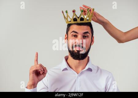 Porträt eines lächelnden bärtigen Geschäftsmanns mit einer goldenen Krone auf dem Kopf auf weißem Hintergrund, aufgenommen im Studio Stockfoto