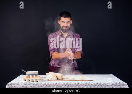 Der junge und attraktive kaukasische Bäcker mit Bart macht den Teig mit Mehl auf schwarzem Hintergrund im Atelier bestreut. Konditor backt Kuchen Stockfoto