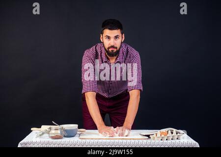 Der junge und attraktive italienische Bäcker mit Bart macht den Teig mit Mehl bestreut italienische Pizza auf schwarzem Hintergrund im Studio Stockfoto