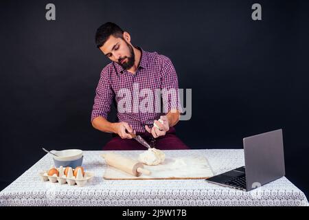 Der junge und attraktive kaukasische Bäcker mit Bart macht den Teig mit Mehl auf schwarzem Hintergrund im Atelier bestreut. Konditor backt Kuchen Stockfoto