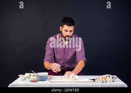 Der junge und attraktive italienische Bäcker mit Bart macht den Teig mit Mehl bestreut italienische Pizza auf schwarzem Hintergrund im Studio Stockfoto