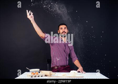 Der junge und attraktive kaukasische Bäcker mit Bart macht den Teig mit Mehl auf schwarzem Hintergrund im Atelier bestreut. Konditor backt Kuchen Stockfoto