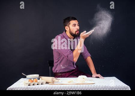 Der junge und attraktive kaukasische Bäcker mit Bart macht den Teig mit Mehl auf schwarzem Hintergrund im Atelier bestreut. Konditor backt Kuchen Stockfoto