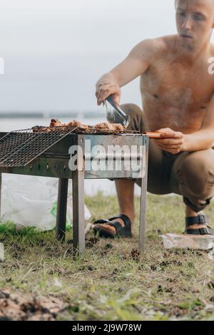 Porträt eines lächelnden Mannes, der Fleisch auf dem Grill würzt. Hübscher Mann, der Grill zubereitet und Rindfleisch auf der Grillparty im Freien für einen Freund zubereitet. Nahaufnahme Hand gri Stockfoto