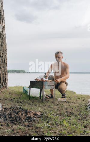 Porträt eines lächelnden Mannes, der Fleisch auf dem Grill würzt. Hübscher Mann, der Grill zubereitet und Rindfleisch auf der Grillparty im Freien für einen Freund zubereitet. Nahaufnahme Hand gri Stockfoto