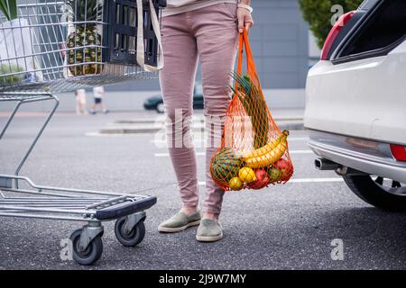 Wiederverwendbarer Netzbeutel mit gekauften Lebensmitteln. Frau, die auf dem Parkplatz steht, nachdem sie im Supermarkt plastisch frei einkaufen konnte. Ethischer Konsum und Nachhaltigkeit Stockfoto