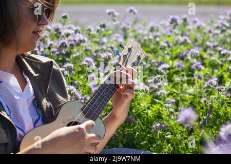 Glückliche Frau, die auf der Ukulele auf der blühenden Wiese spielt. Musikgenuss und Frühling im Freien Stockfoto