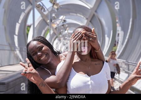 Zwei afroamerikanische Frauen bedeckten ihre Augen und umarmten sich von hinten. Zwei überraschte Mädchen. Frauen, die Spaß haben und Gesichtsemotionen zeigen Stockfoto